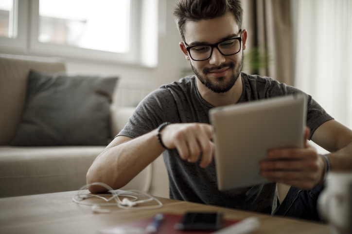 Jovem de cabelos escuros, usando óculos ,mexendo em um dispositivo móvel.